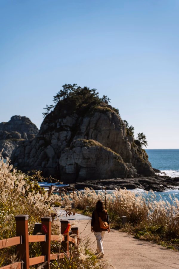 A woman walks along a path on Jeju Island with a large rock monolith rising from the sea in the background