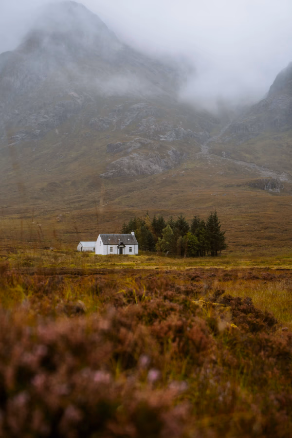 A small white cottage known as the Wee White House sits in a green valley in front of the misty mountains of Glencoe, Scotland.