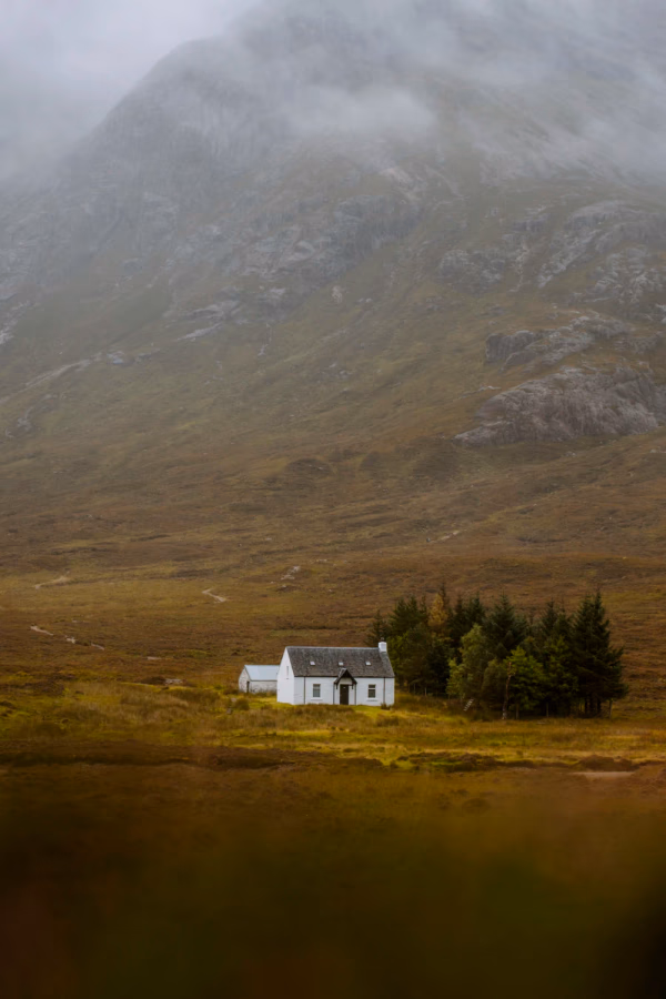 A small white cottage sits across a calm and misty loch