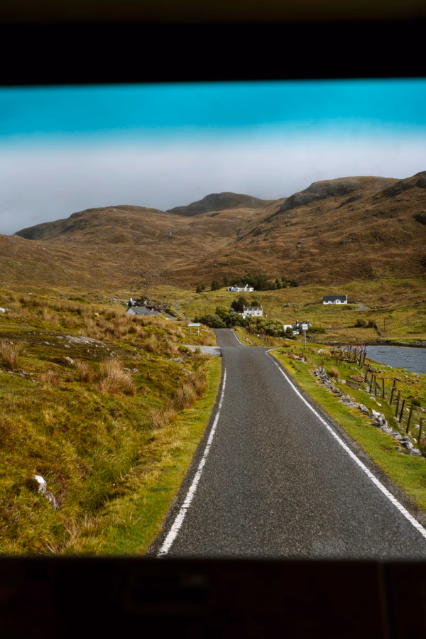 A single track road framed by the windshield of a vehicle leads to several white cottages