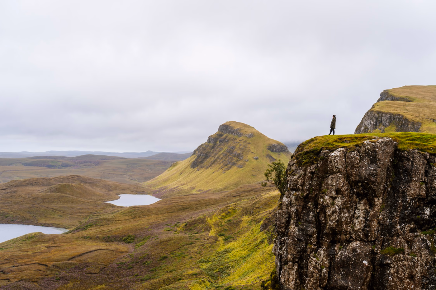 A man stands in the distance at the edge of a cliff looking down at the valley below on The Isle of Skye, Scotland