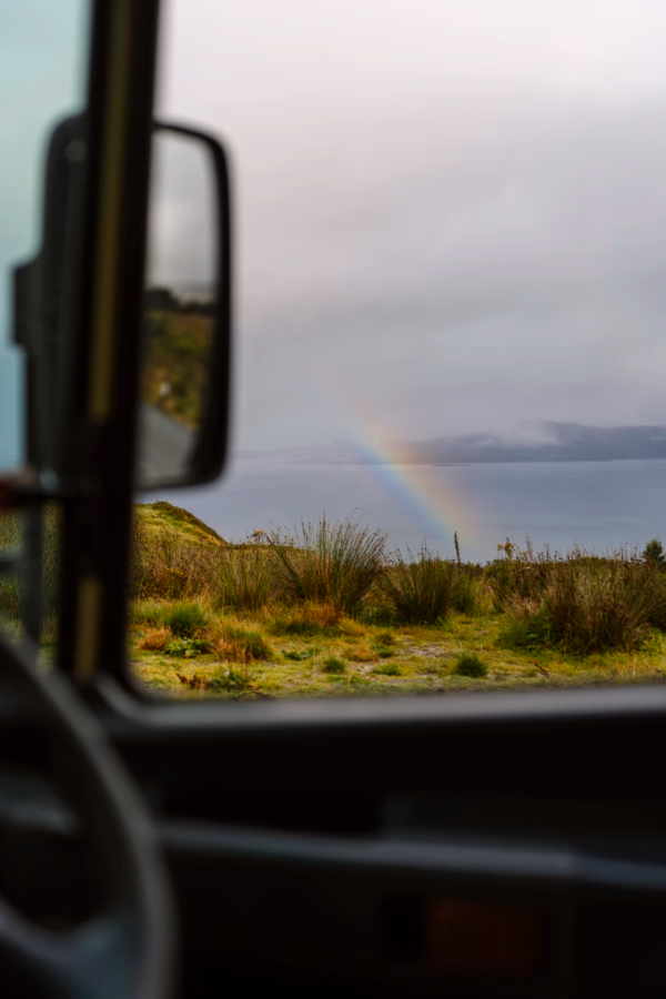 A rainbow streaks across the sky through the side window of a vehicle on The Isle of Skye, Scotland