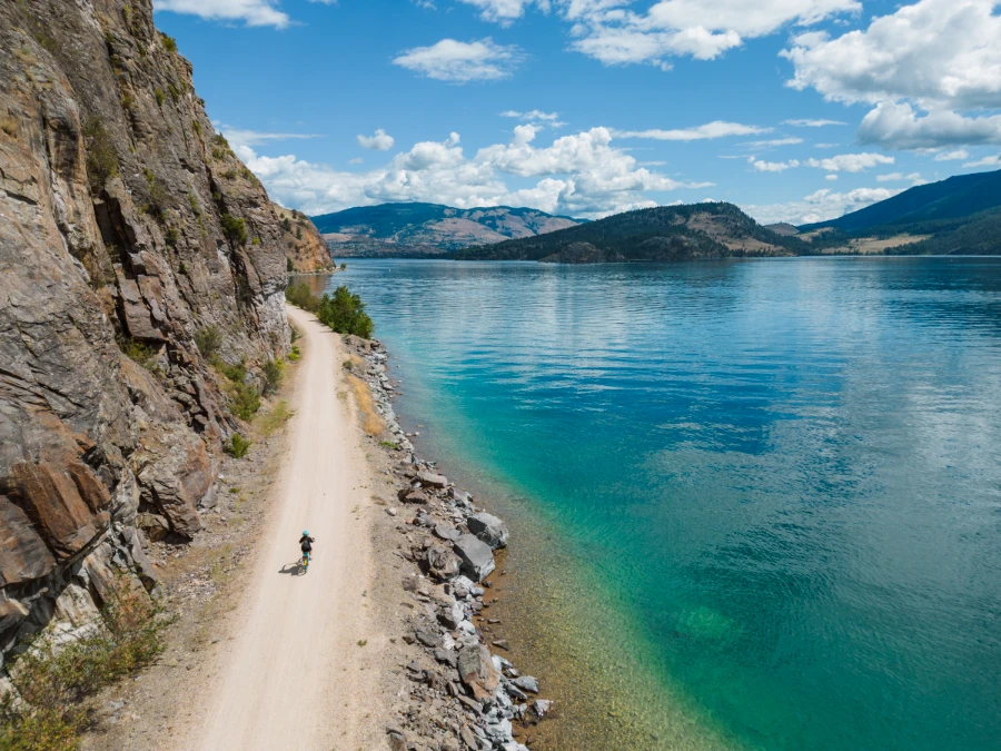 A cyclist rides down the centre of a wide dirt path with a steep stone cliff rising to their left, and turquoise waters to their right