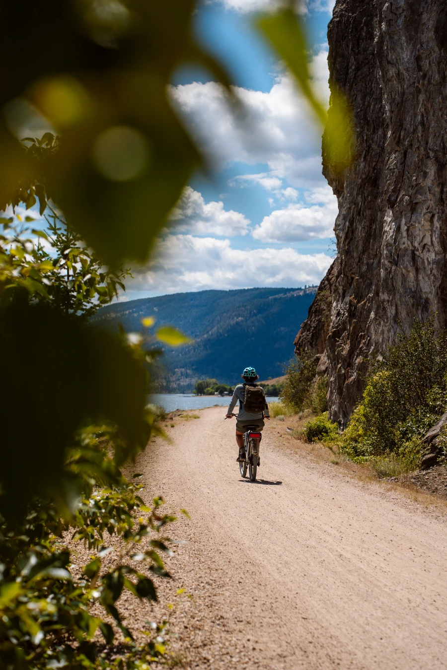 Framed by a bush of green leaves, a cyclist rides down a curved flat gravel path with a steep stone cliff to their right