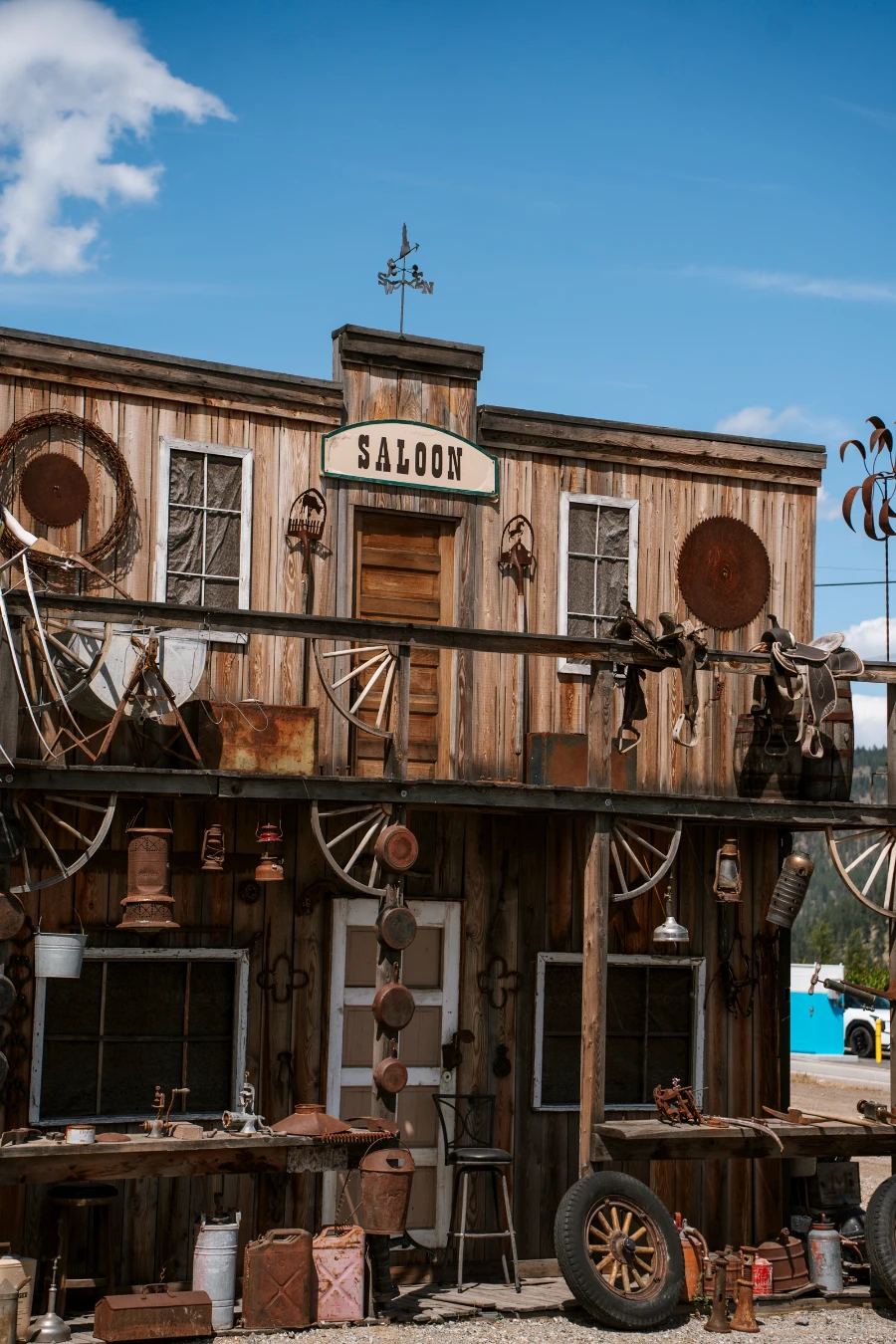 A frontier-era-style wooden building is cluttered with wooden wheels, old gas canisters, and tin pots hanging from its facade and has a sign in the centre that reads ‘Saloon’