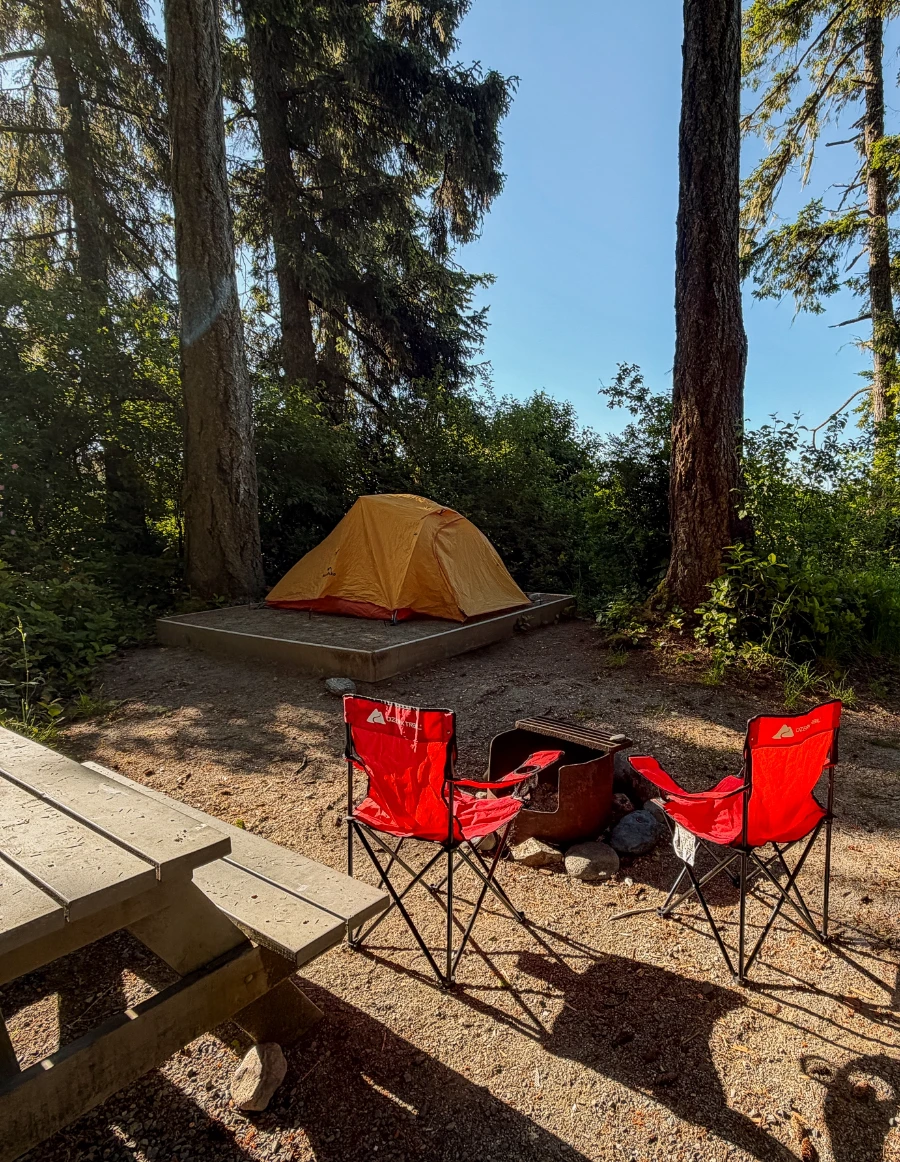 Two red camp chairs set up next to a picnic table and a fire pit, with an orange tent set up on a tent pad in the distance