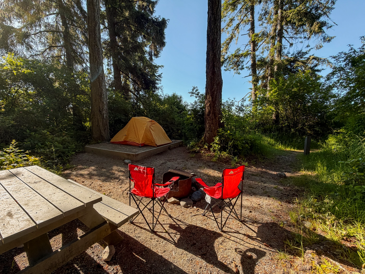 Two red camp chairs and a wooden picnic table set up at a dirt camp site next to a platform where a red tent is set up surrounded by trees near the coast in Jordan River