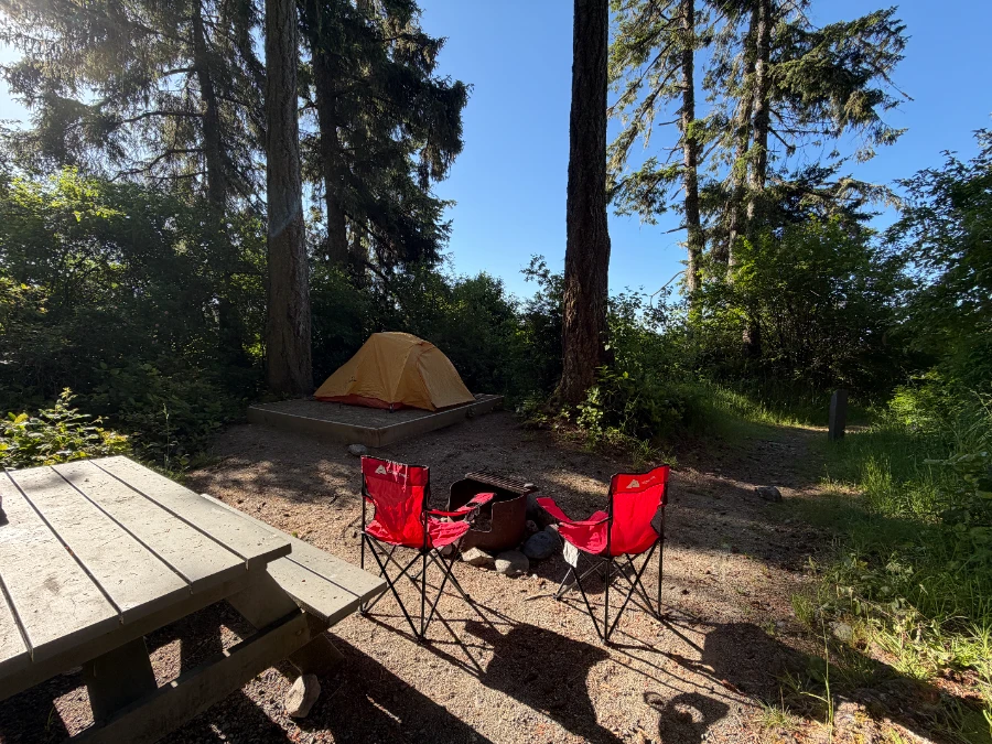 Two red camp chairs and a wooden picnic table set up at a dirt camp site next to a platform where a red tent is set up surrounded by trees near the coast in Jordan River