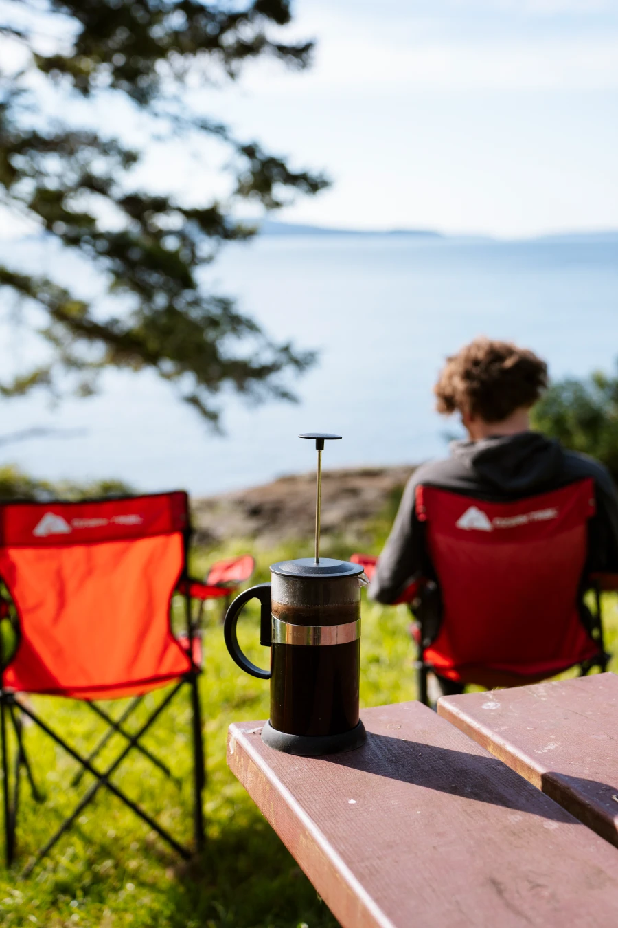 A french press full of coffee sits on a picnic table next to two red camp chairs