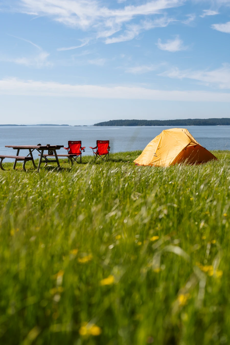 Two red camp chairs, an orange tent, and a wooden picnic table sit on a grassy field next to the coast at Ruckle Provincial Park