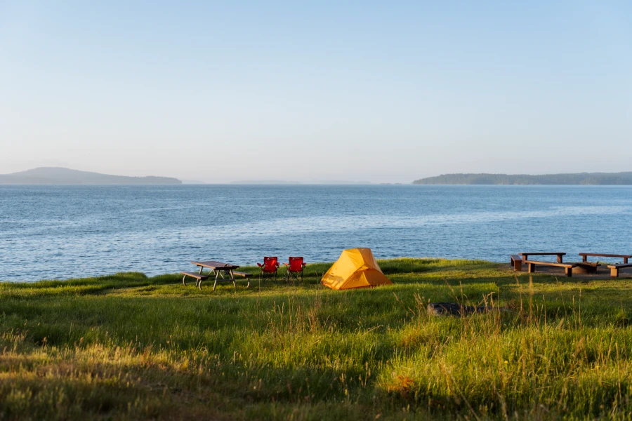 A field of grass next to the coast with a picnic table, camp chairs, and tent set up