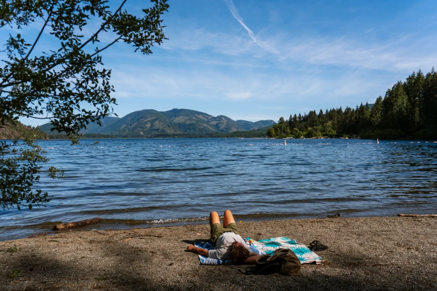 A man lying on a beach towel on the sandy bank along the edge of Cowichan Lake
