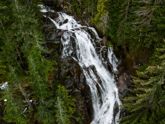 Powerful stream of water pours over a polished granite cliff and makes a sharp 90 degree turn, surrounded by lush green trees