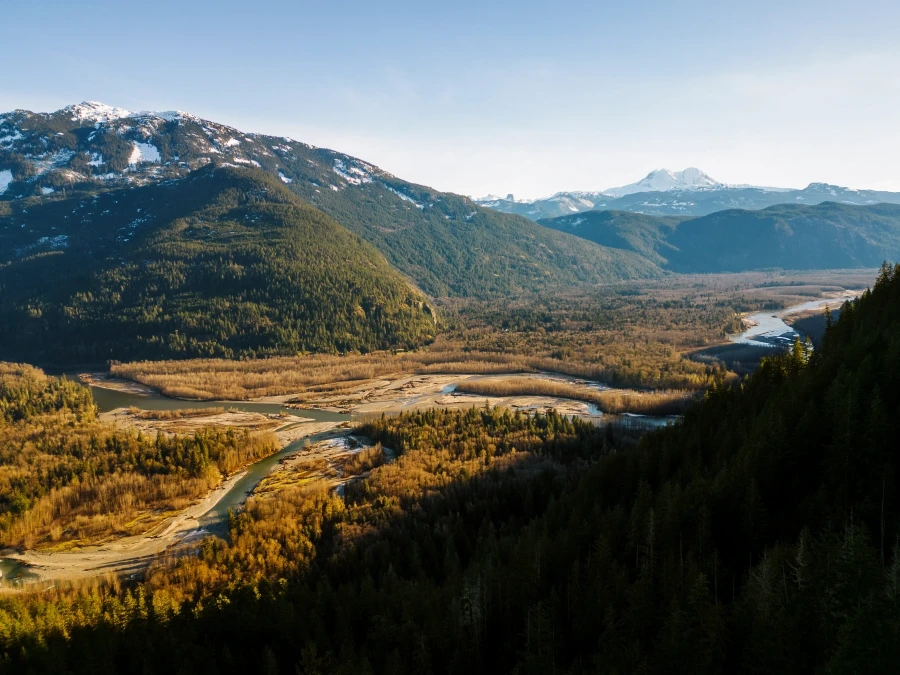 A meandering river flows through a valley of dry trees next to rolling foothills with larger mountains beyond