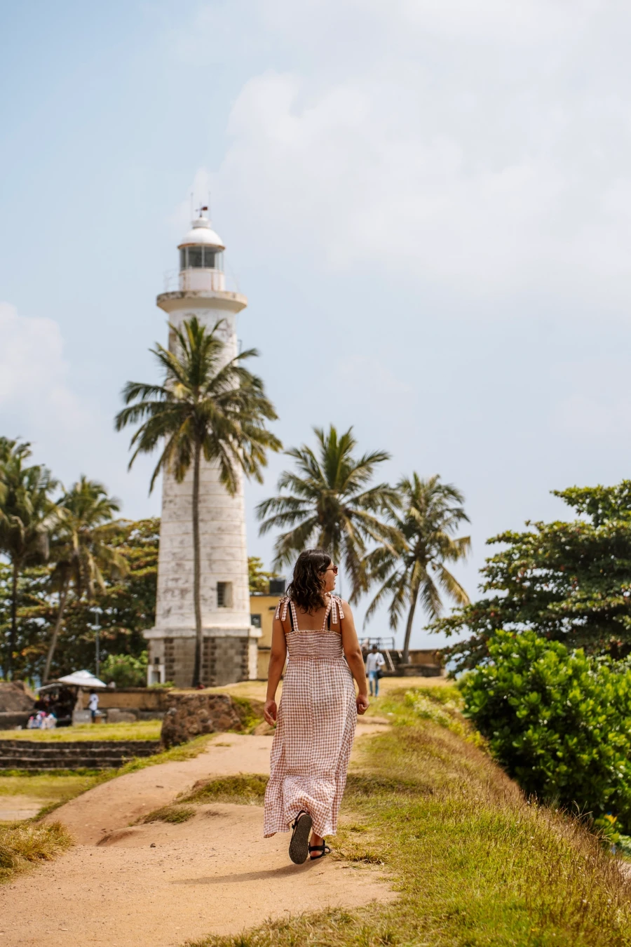 A woman in a summer dress walks along a palm-tree lined path towards a white stone tower in Galle, Sri Lanka