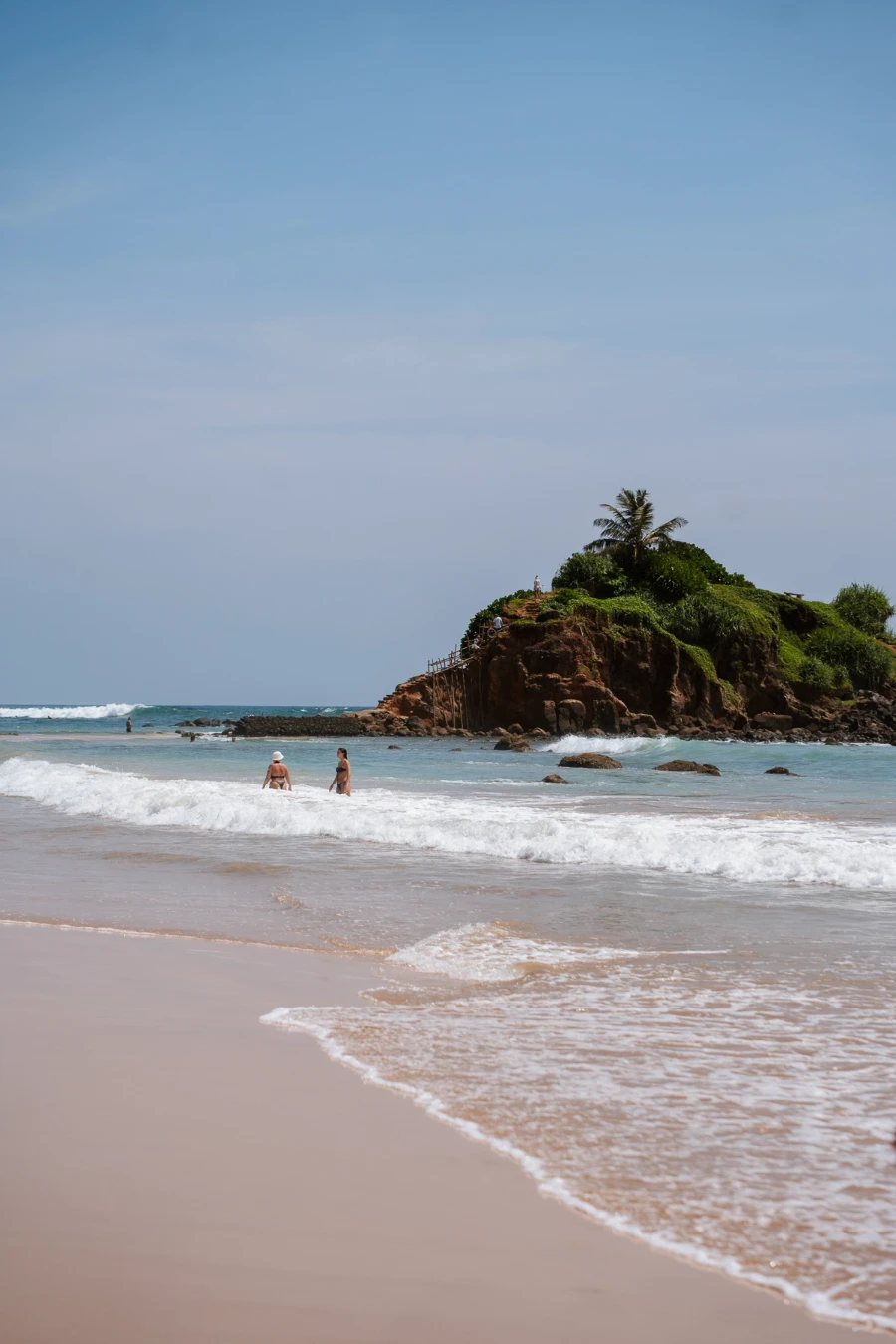 Choppy white-wash waves crash onto the flat sandy shores of Mirissa beach while two people stand knee deep in the water