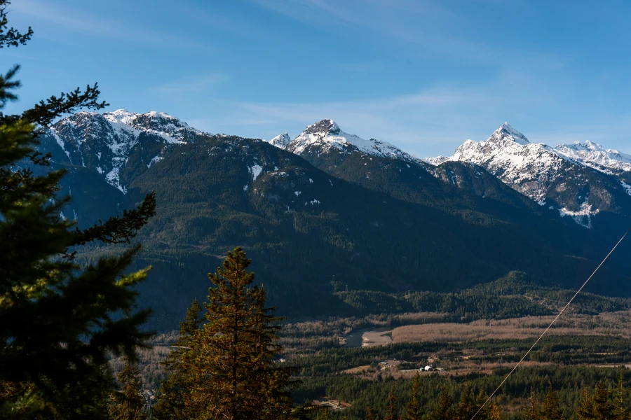 A dry valley with a river running through it has several forested mountains with snowy peaks bordering it