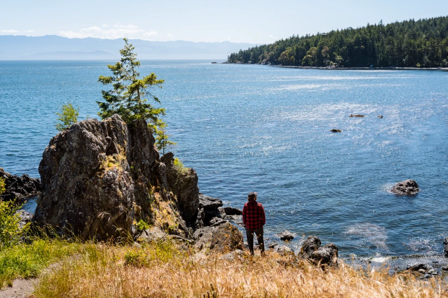 A man stands at the edge of a cliff above a sparkling calm sea of blue water on East Sooke Regional Park