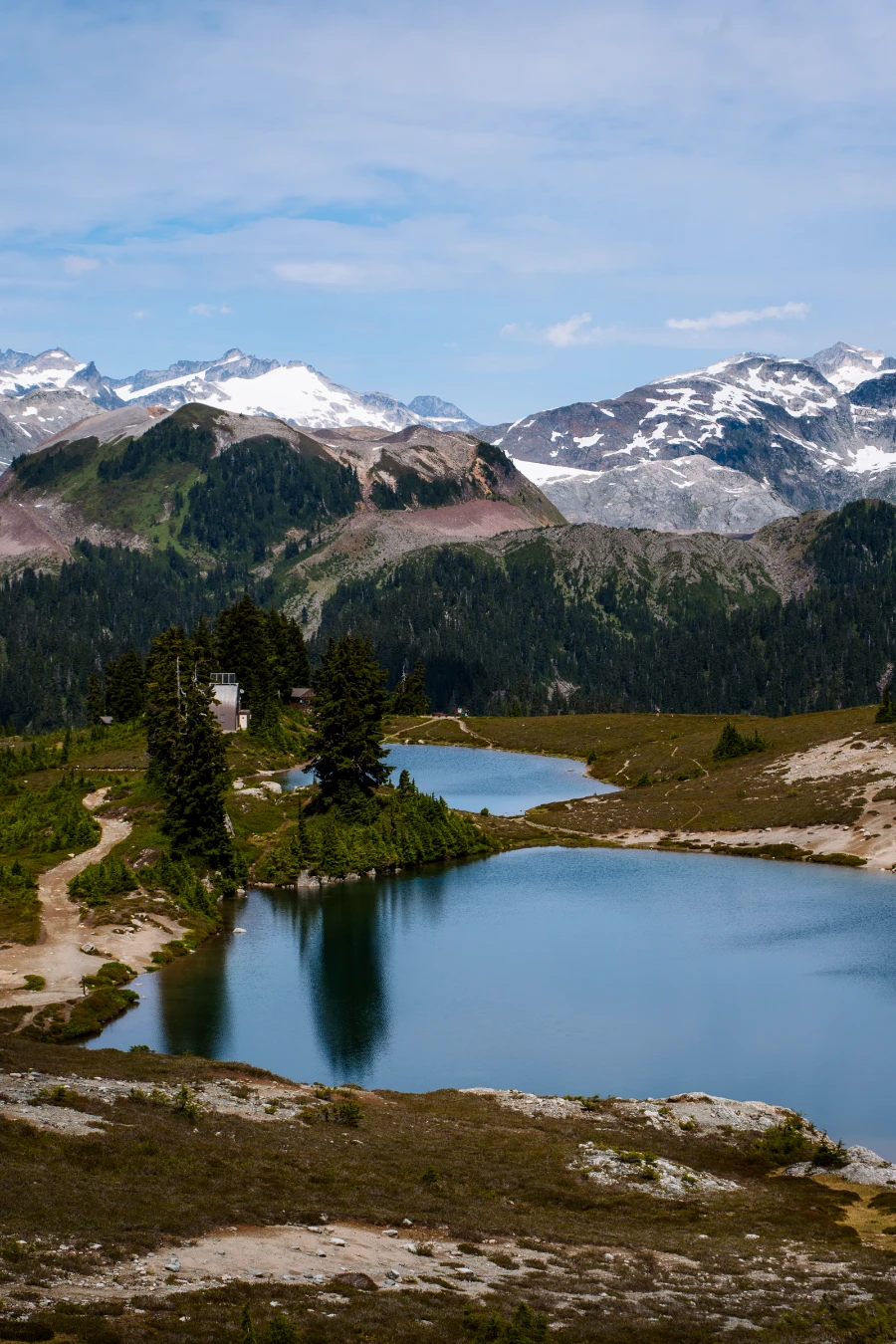 A narrow dirt path runs along the left of Elfin Lakes with several snow-capped peaks in the distance