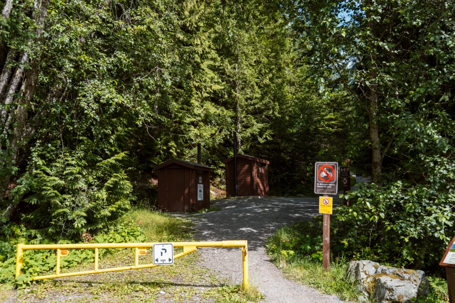 A yellow gate is shut next to a narrow gravel trail that leads into the woods, with pit toilets off the side of the trail.