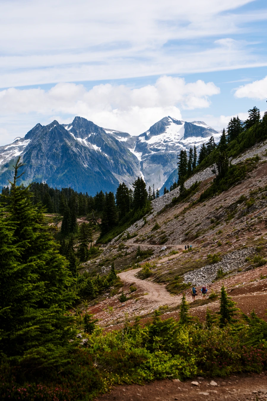 A dirt path winds along a mountain ridge with snow-covered peaks in the distance