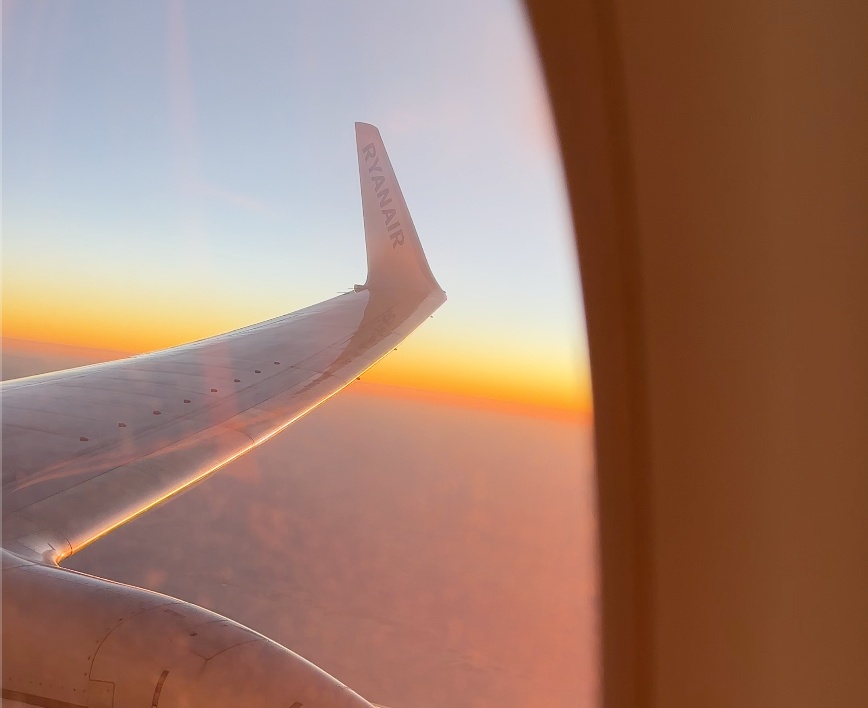 The wing of an airplane seen through a passenger’s window at sunset while the plane flies above the cloud layer