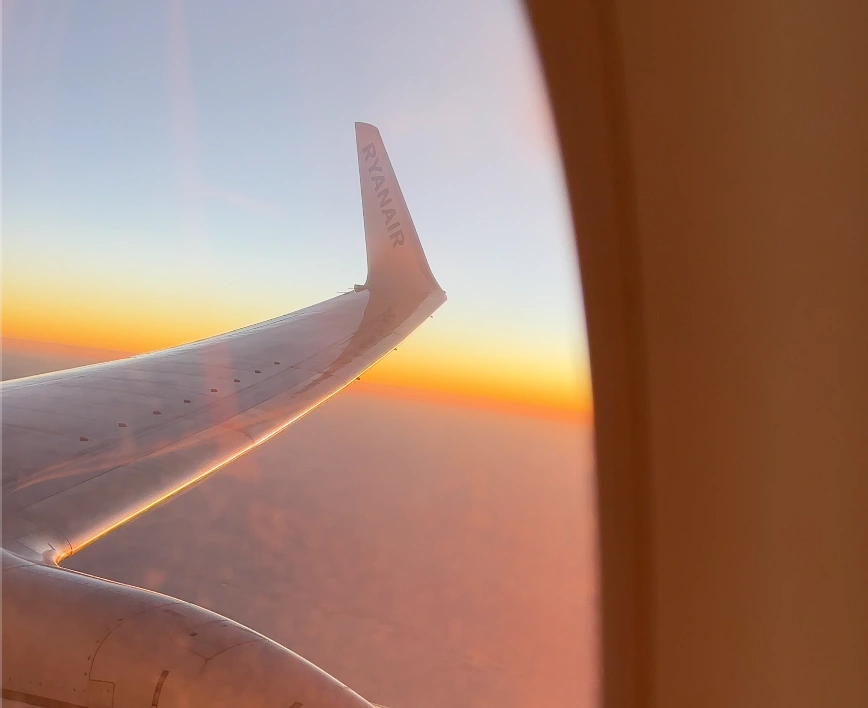 The wing of an airplane seen through a passenger’s window at sunset while the plane flies above the cloud layer