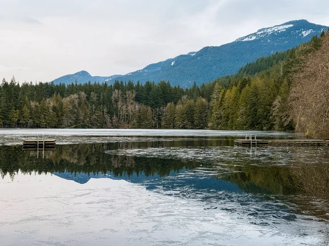 A half frozen lake with a wooden dock is surrounded by ever green trees with mountains in the distance