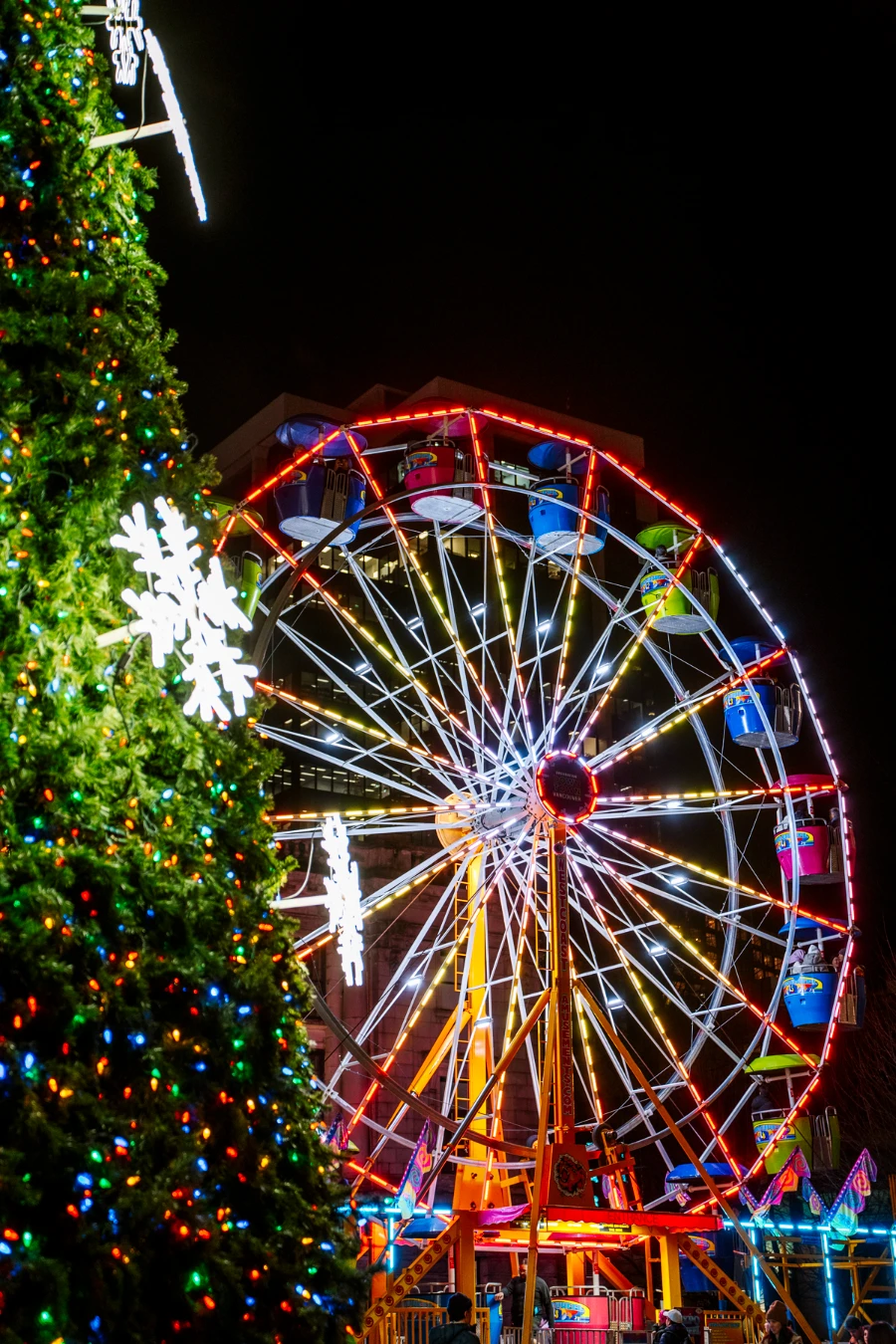 A large ferris wheel is lit several different colors next to a large christmas tree