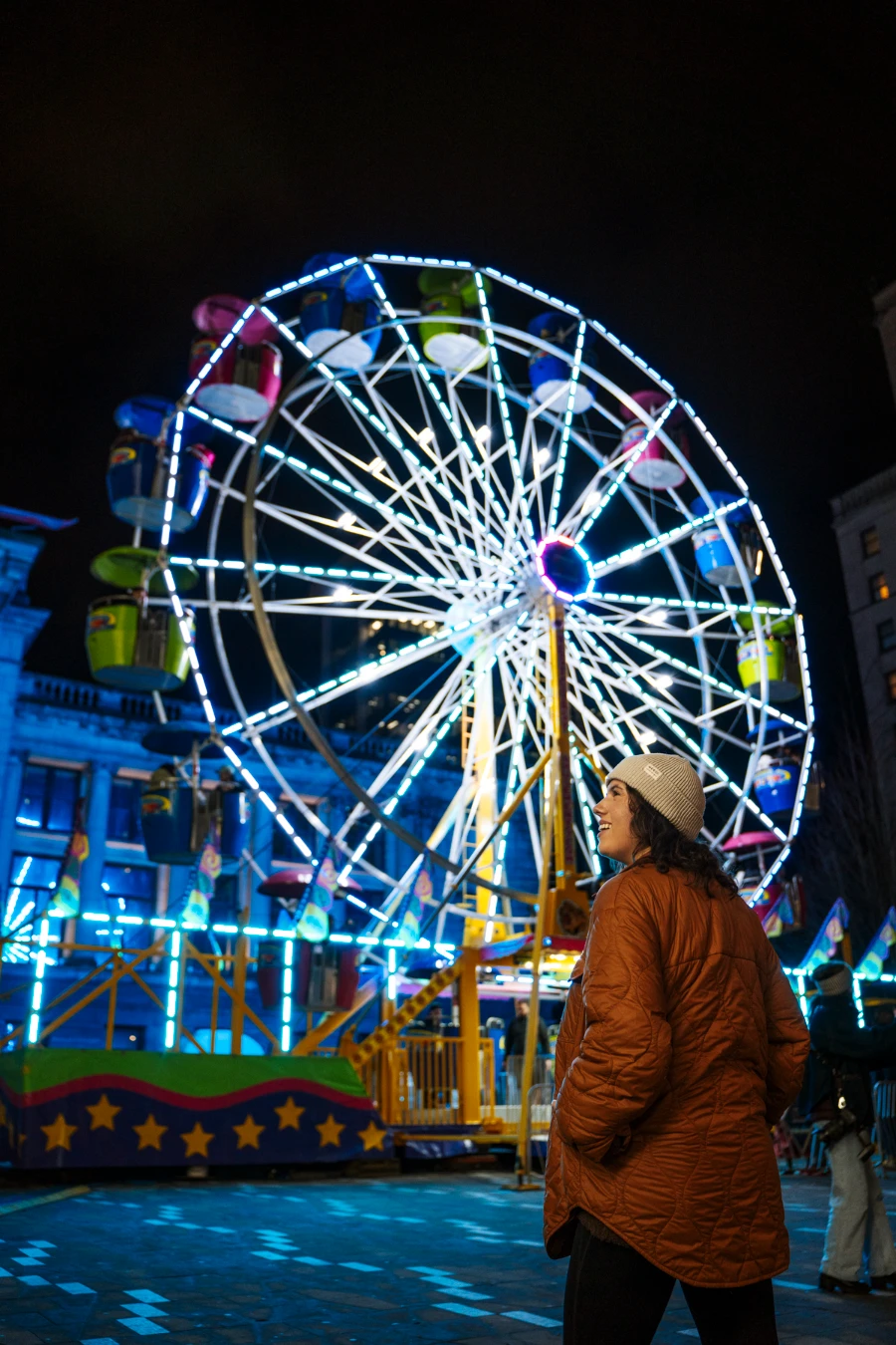 A woman in winter attire stands in front of a lit up ferris wheel at night in downtown Vancouver