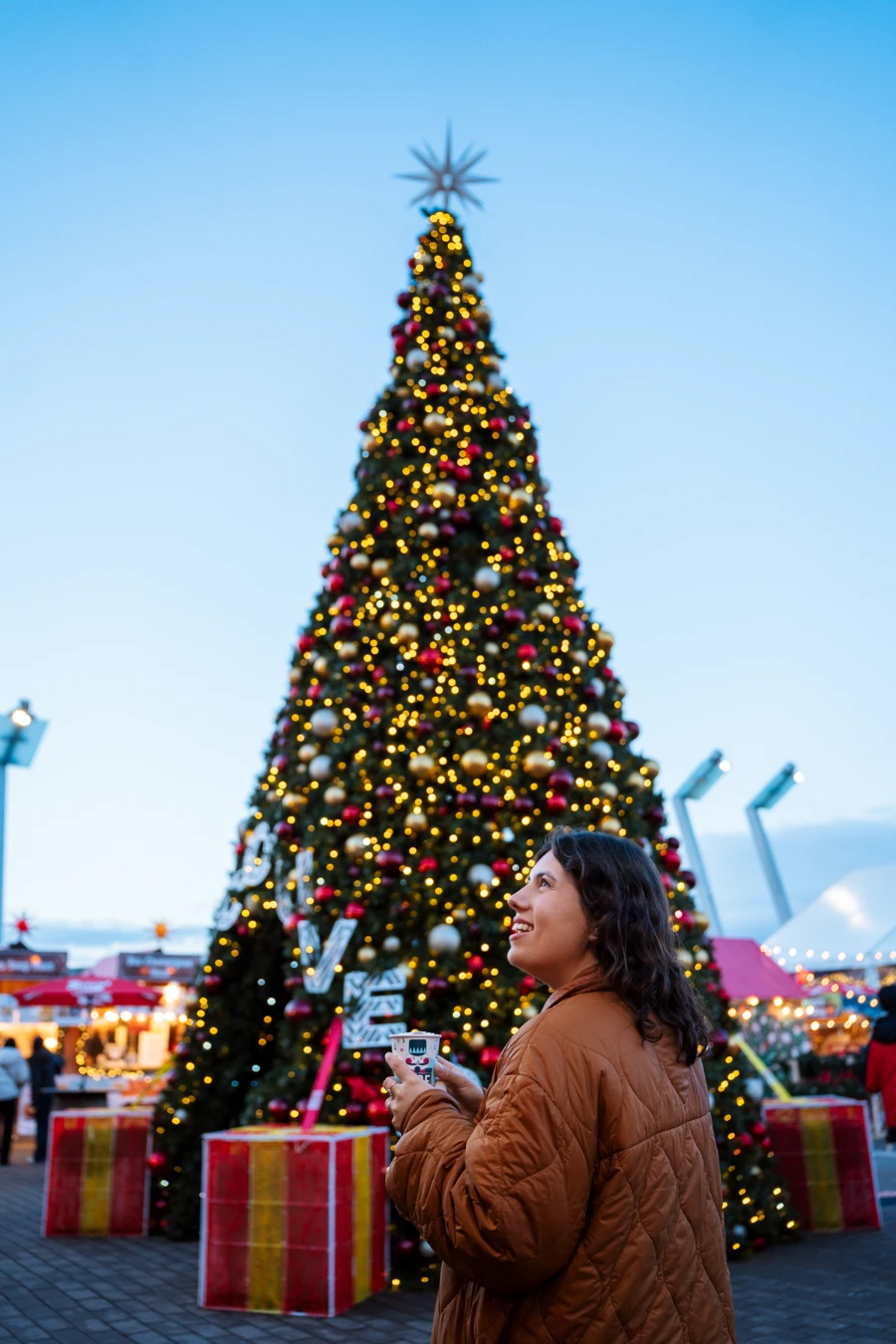 A woman holds a hot chocolate in front of a large decorated Christmas tree at the Downtown Vancouver Christmas market