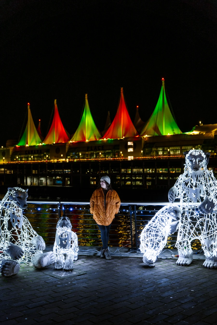 A woman stands next to several string light statues of polar bears with Canada Place in the distance behind her