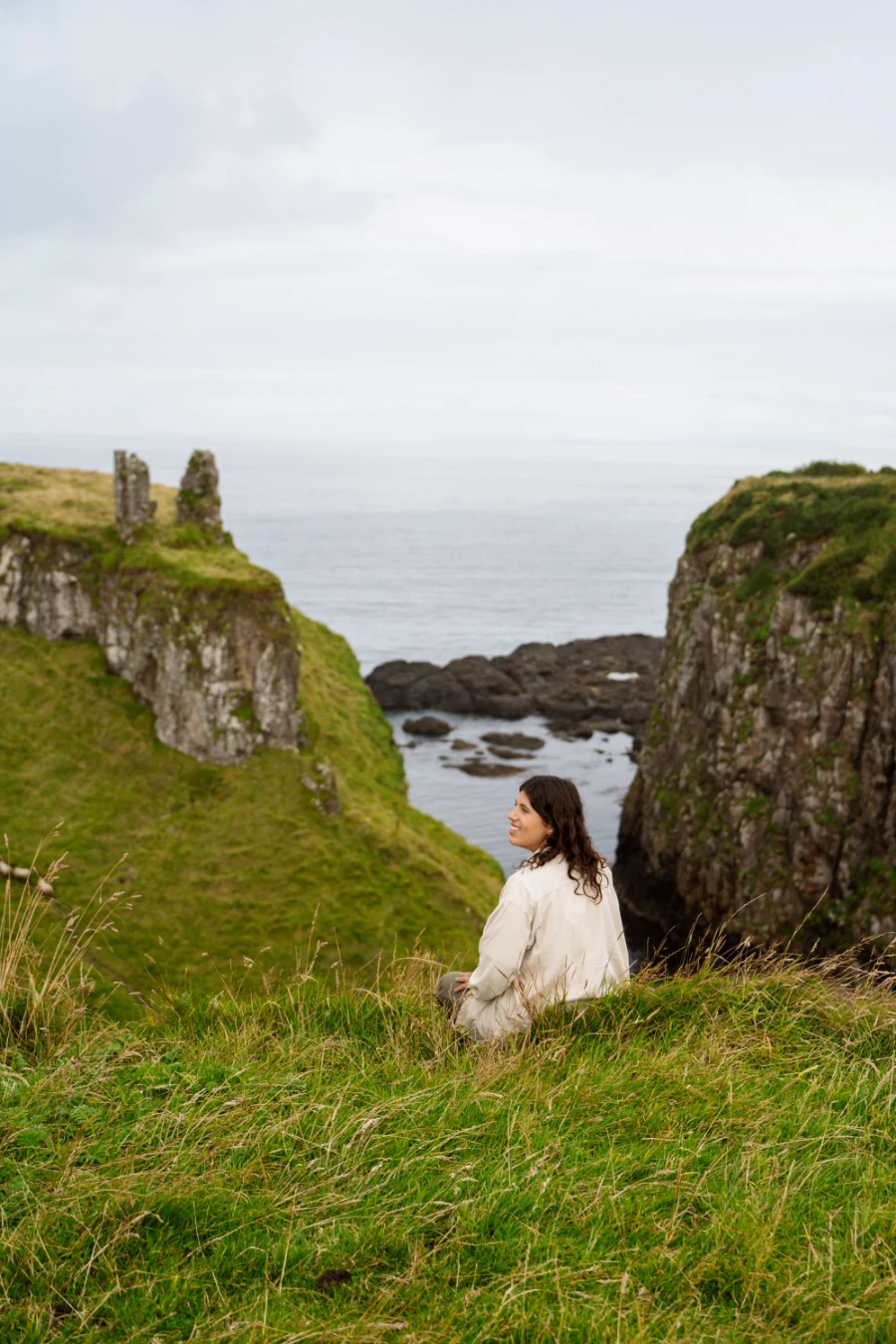 A woman sits on a grassy cliff looking down at a distant grassy valley