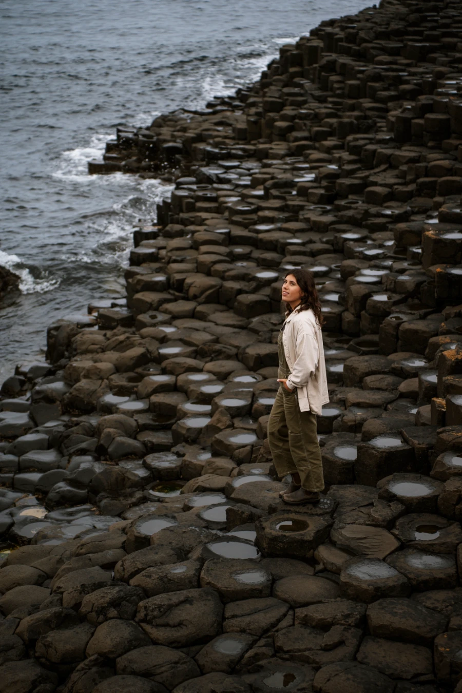 A woman stands on a patch of interlocking haxagon-shaped pillars form an uneven surface and is wet from the spray of the sea