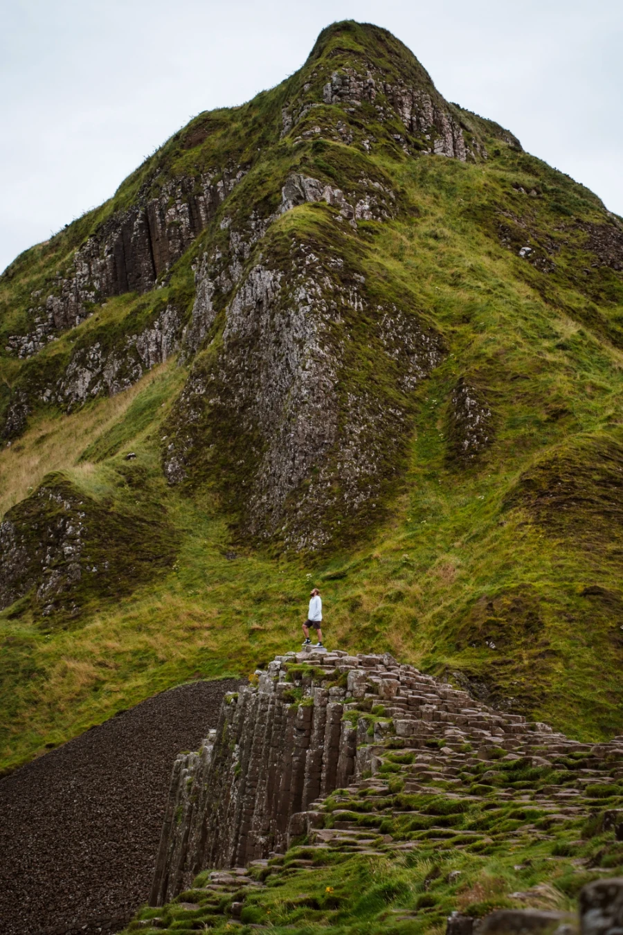 A man stands at the end on a rocky outcropt comprised of hexagon-shaped pillars with a steep grassy mountain behind him