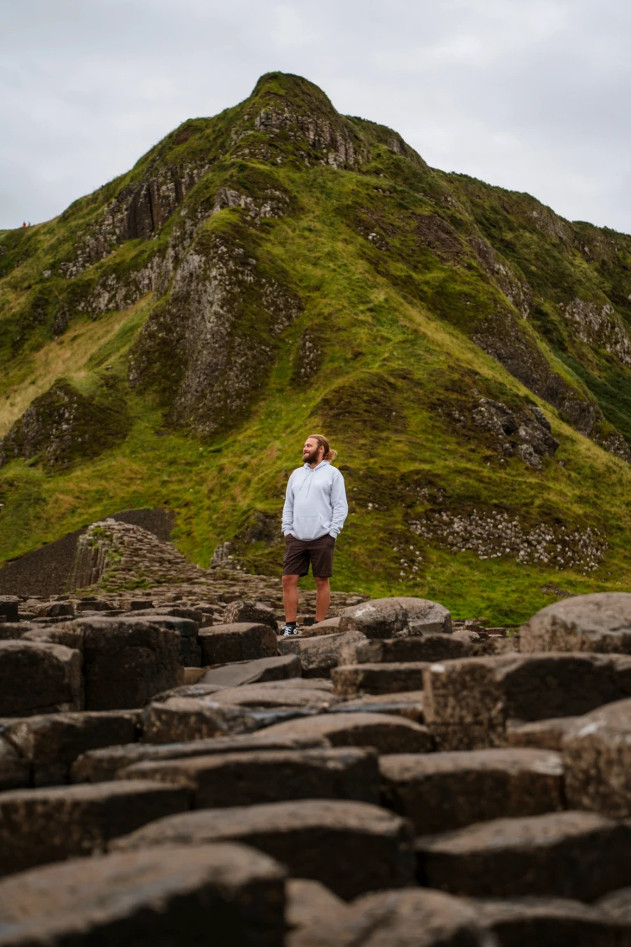 A man standing on a stretch of interlocking haxagon-shaped pillars in front of a steep grassy mountain