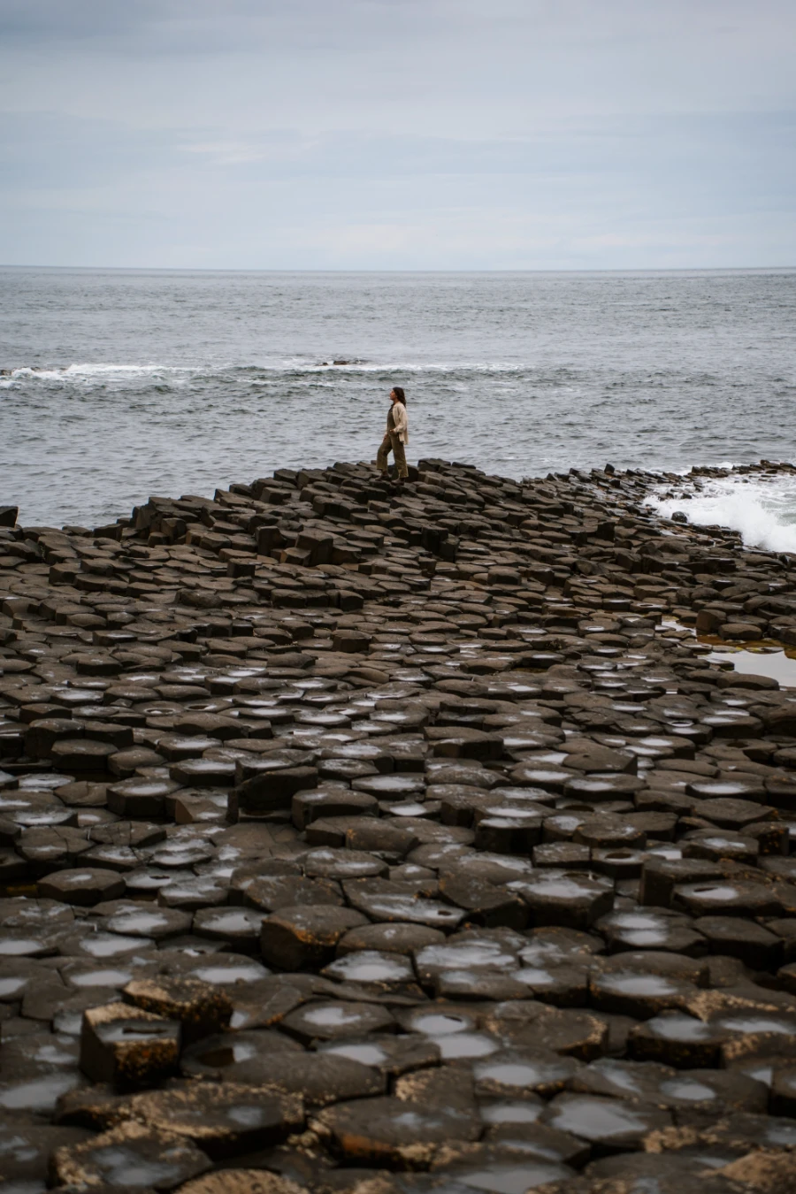 A woman stands at the end of a long stretch of interlocking haxagon-shaped pillars that form an uneven surface and is wet from the spray of the sea
