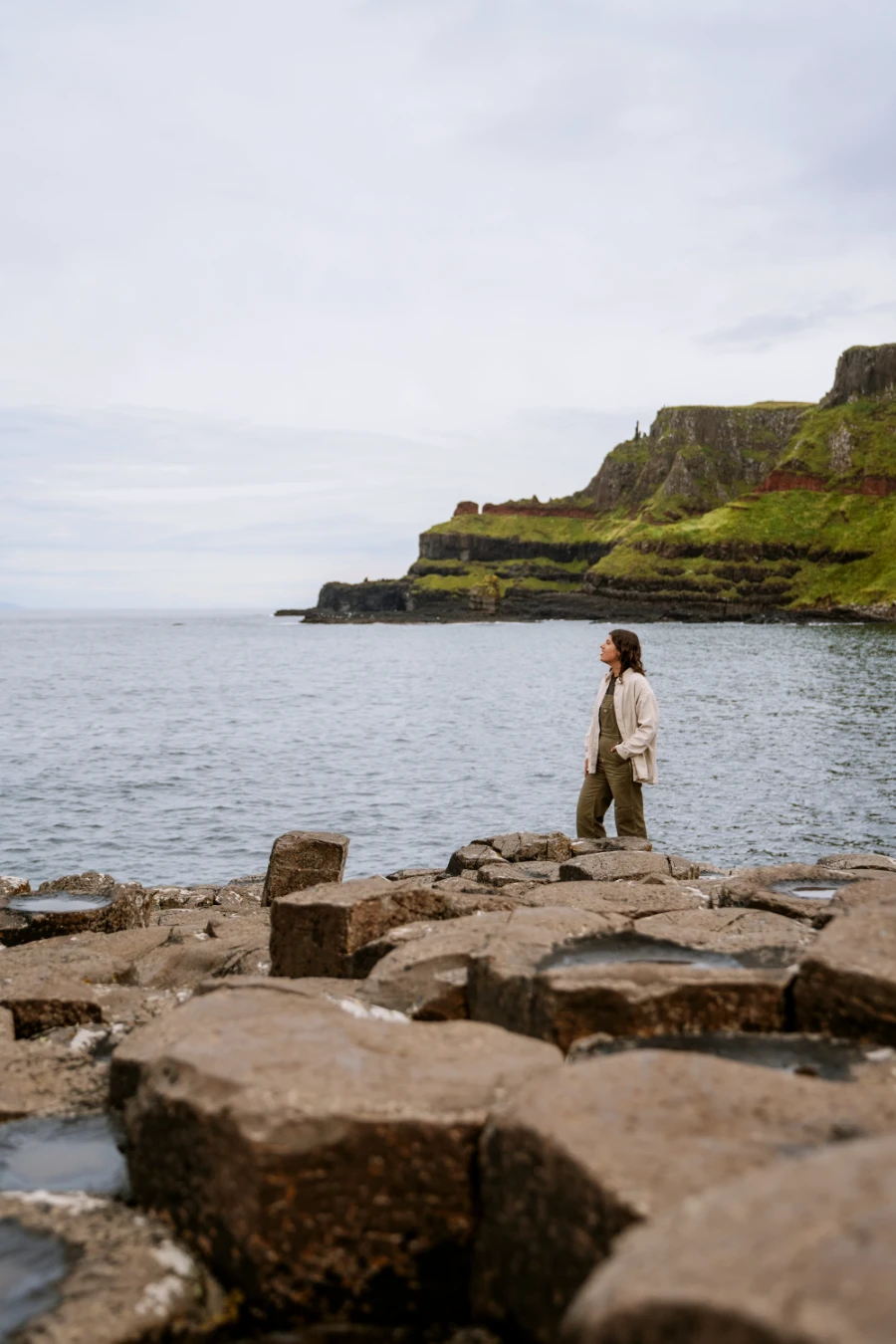 A woman stands on the rocks of The Giant's Causeway with a calm sea and steep grassy cliffs behind her