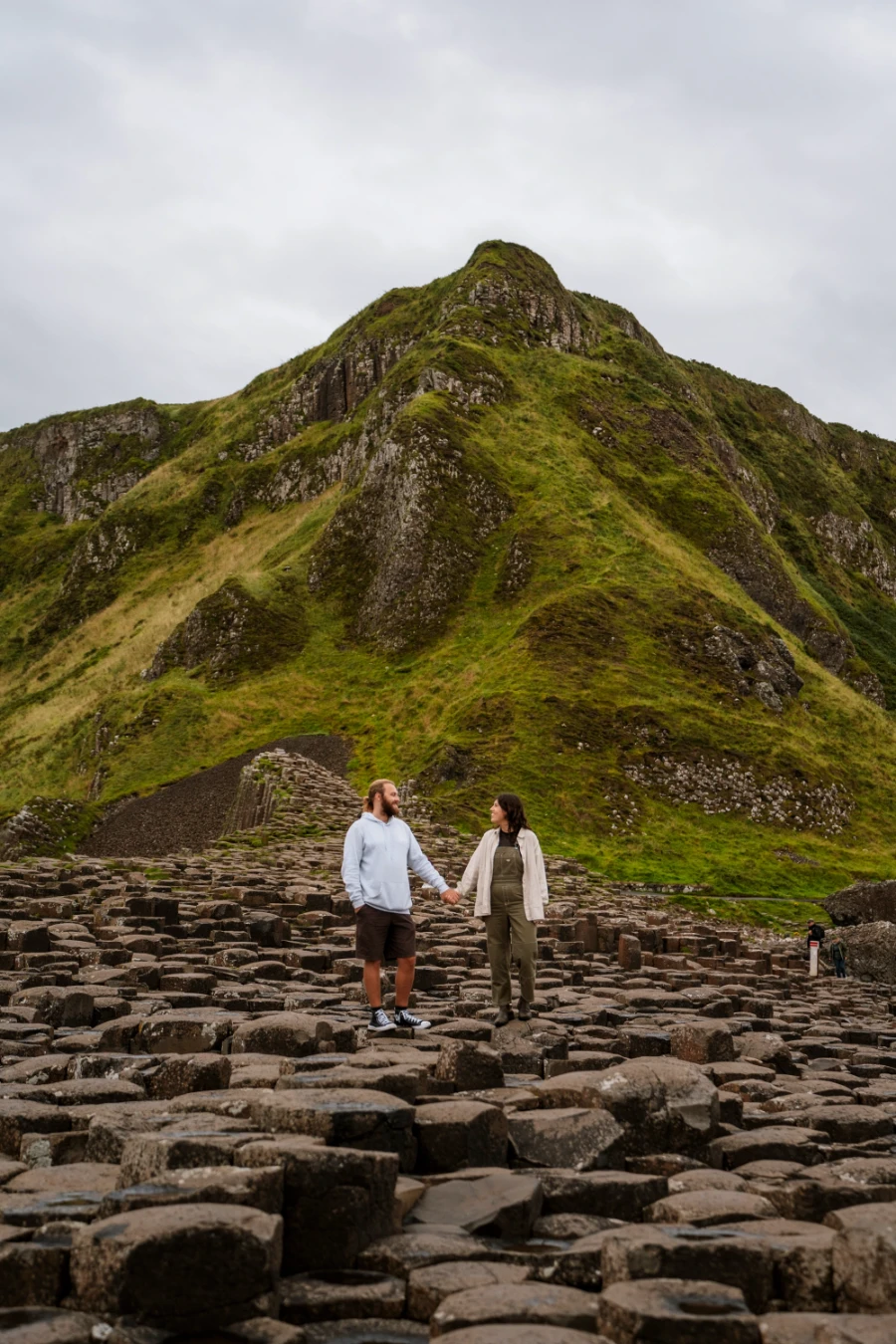 A man and woman hold hands on a stretch of interlocking haxagon-shaped pillars with a large grassy mountain behind them