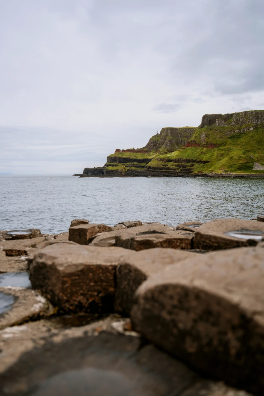 A few layers of interlocking haxagon-shaped pillars frame a view of the calm sea and green cliffs in the distance