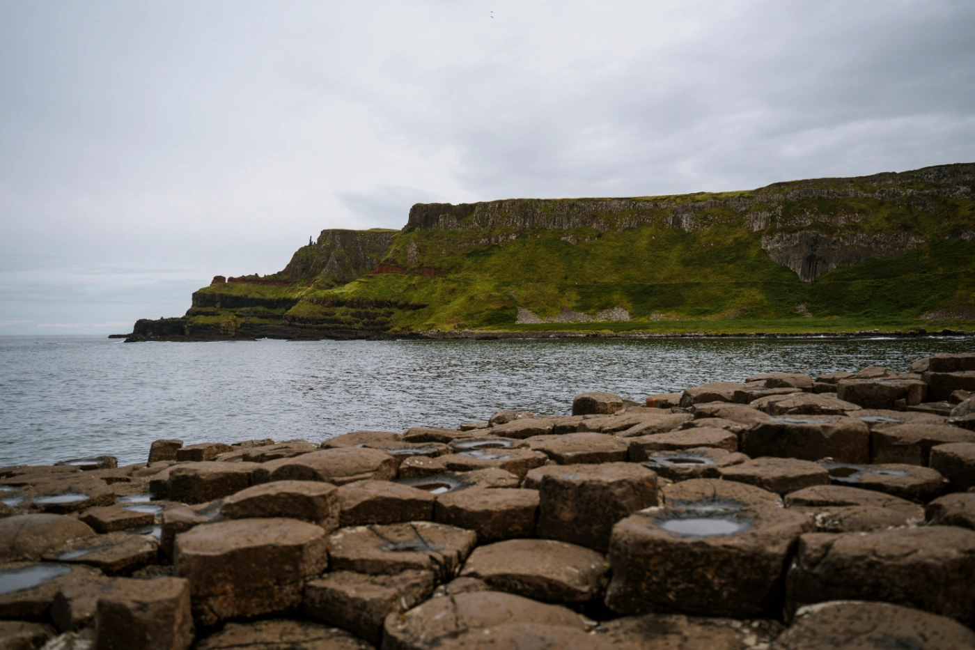 A collection of interlocking haxagon-shaped pillars with the calm sea surrounding them with grassy cliffs in the distance