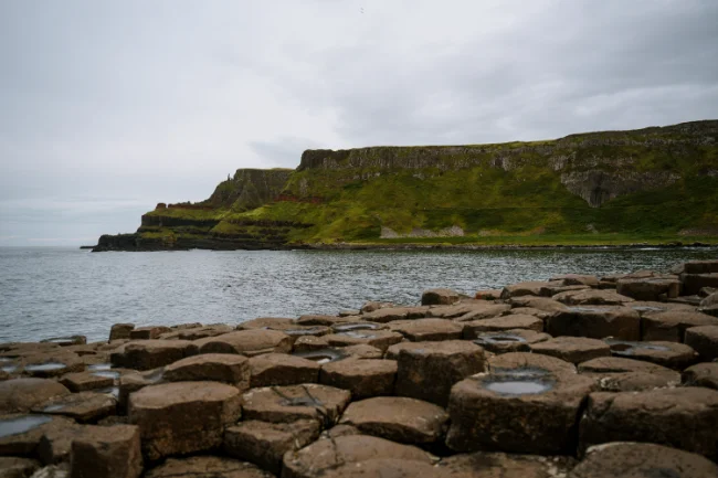 A collection of interlocking haxagon-shaped pillars with the calm sea surrounding them with grassy cliffs in the distance