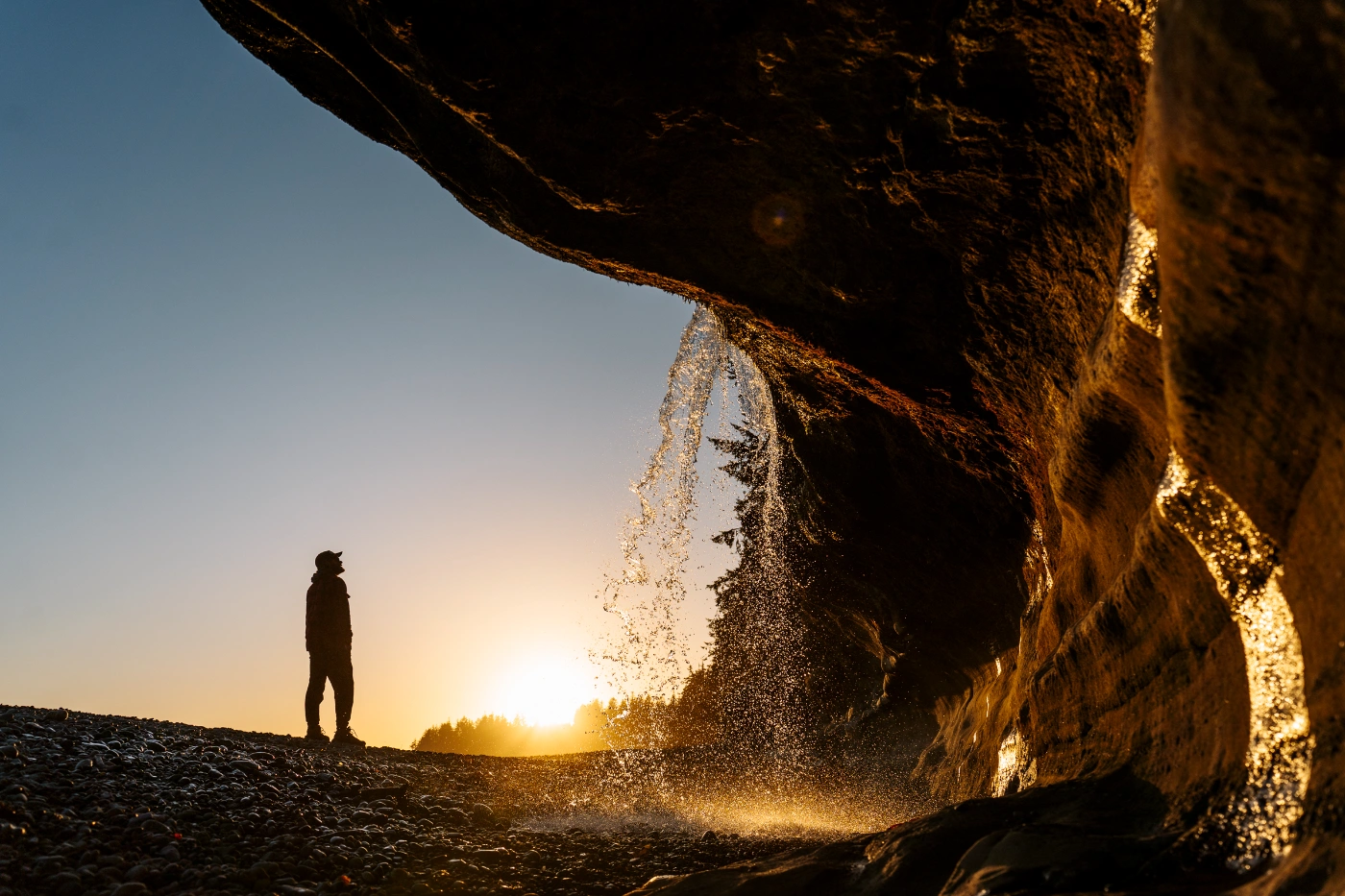 A steady flow of water pours over a sandstone cliff into the rocky beach below while a man walk in the distance with the setting sun behind him