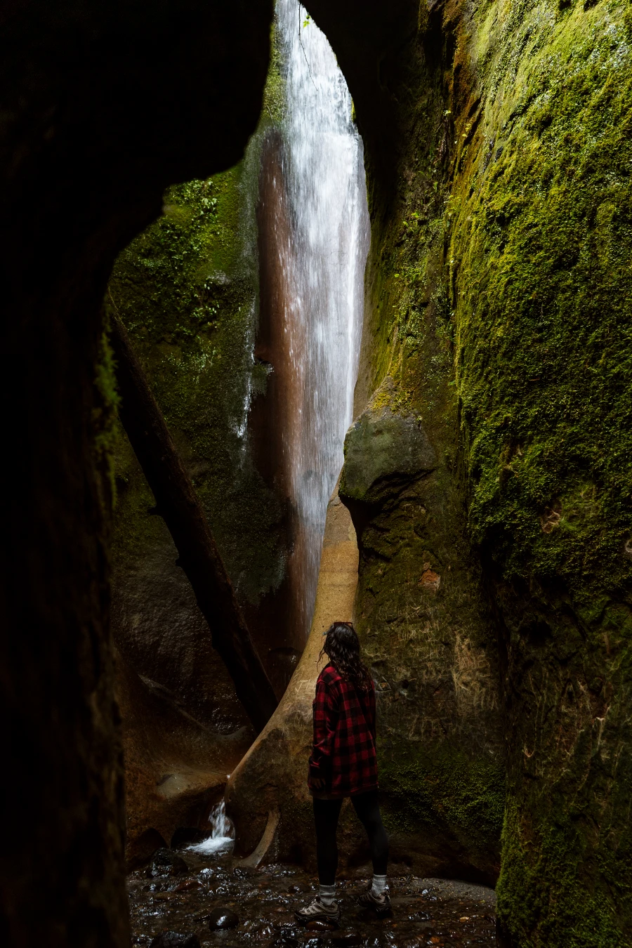 A woman walking through a dark, mossy cavern leads to a spot-lit waterfall at the end