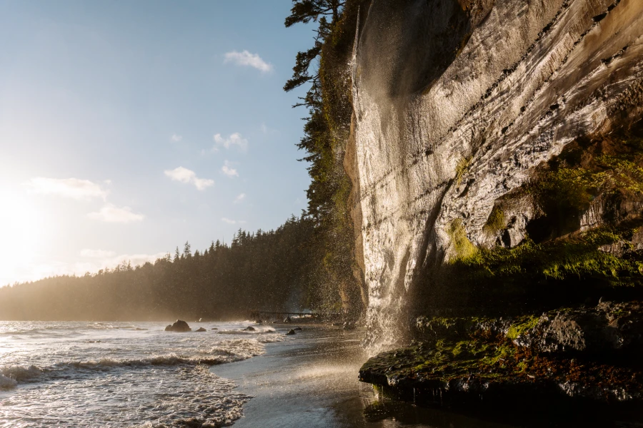 A misty waterfall pours over a cliff into the sea on a sandy beach below at Mystic Beach