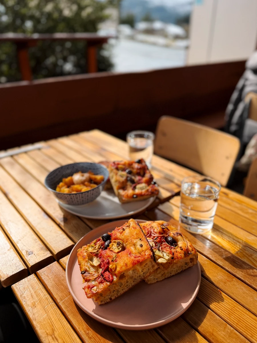 Slice of thick focaccia style pizza are sat on two plates next to glasses of water on a wooden table