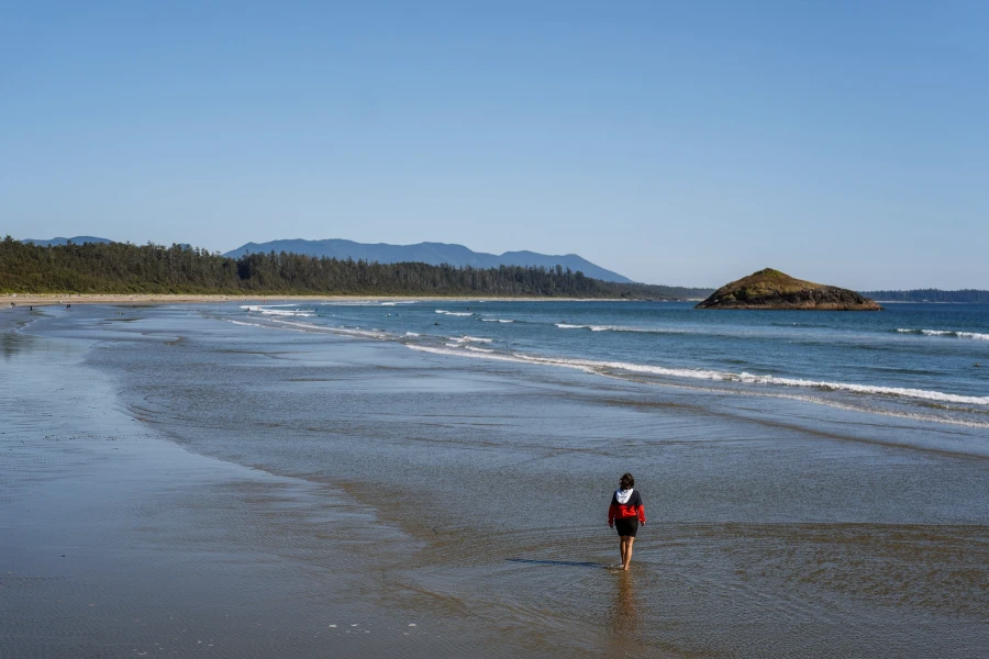 A woman walking in ankle deep water along the flat sandy stretch of Long Beach near Tofino BC while rows of gentle waves approach the shore
