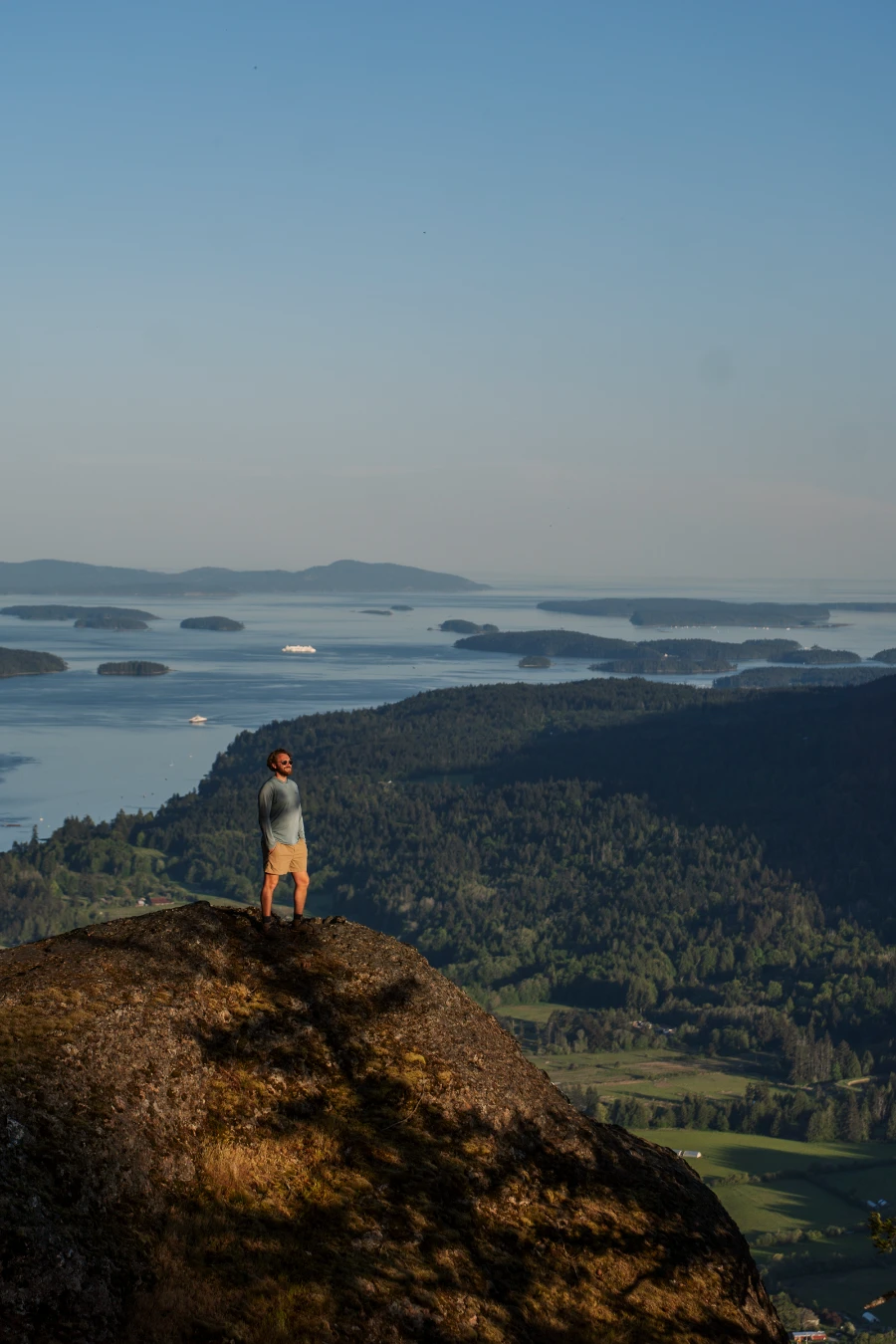 Man stands on a cliffs edge with lush valleys of farmland below him