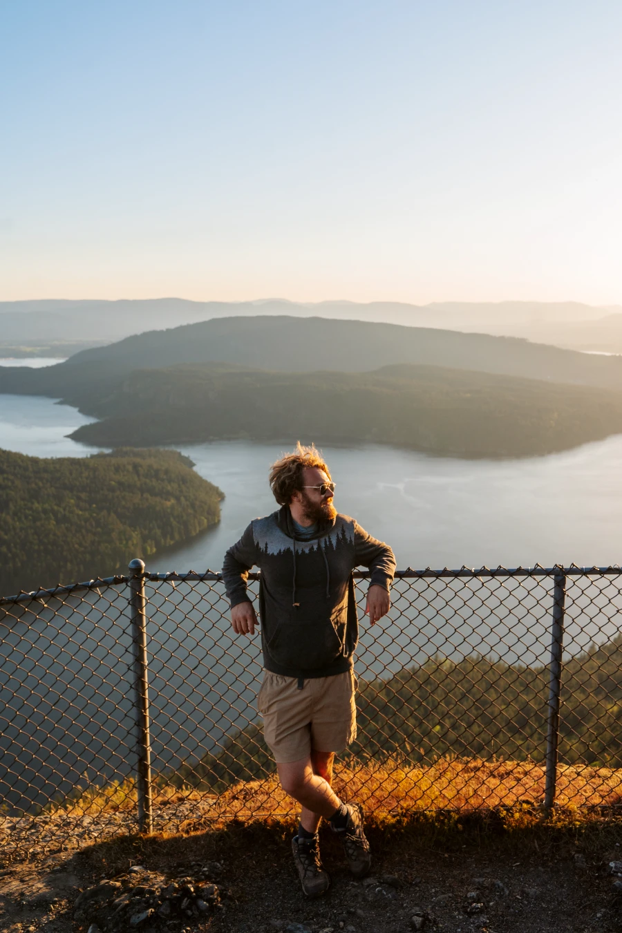 Man leans against a chain link fence on the edge of a cliff above islands sitting in calm seas lit by warm sunset light