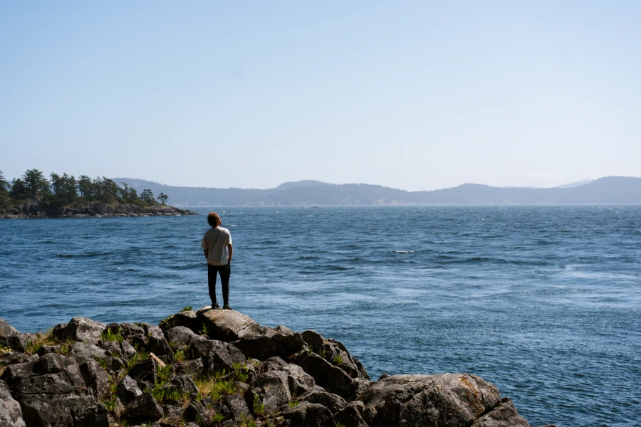 A man stands on a rocky outcrop surrounded by calm blue water with mountains small on the distant horizon